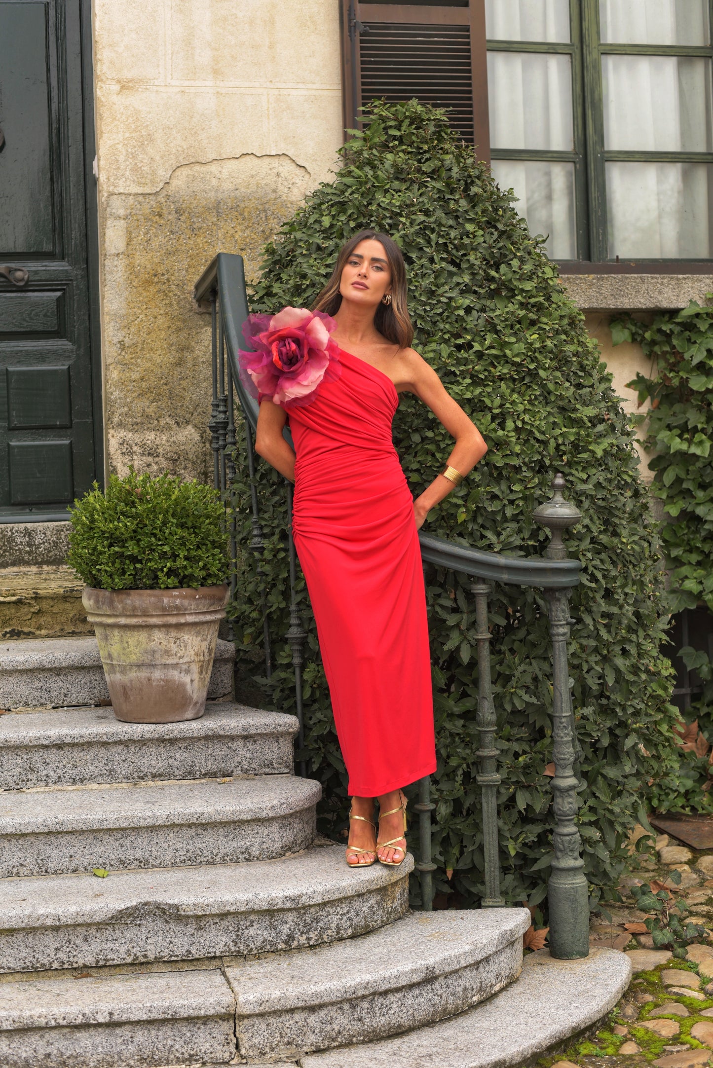 Vestido de Fiesta midi confeccionado en punto de seda color rojo, drapeado con escote asimétrico. Incluye gran broche de flor de organza en tonos fresa que se puede poner o quitar. Vestido forrado. 