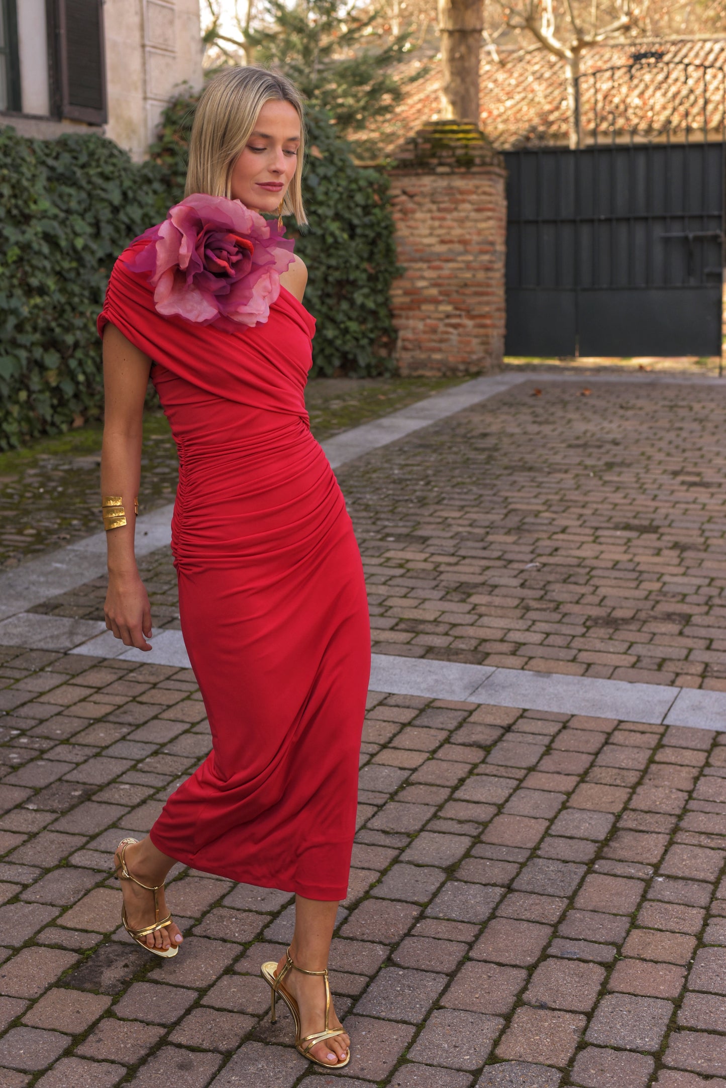 Vestido de Fiesta midi confeccionado en punto de seda color rojo, drapeado con escote asimétrico. Incluye gran broche de flor de organza en tonos fresa que se puede poner o quitar. Vestido forrado. 