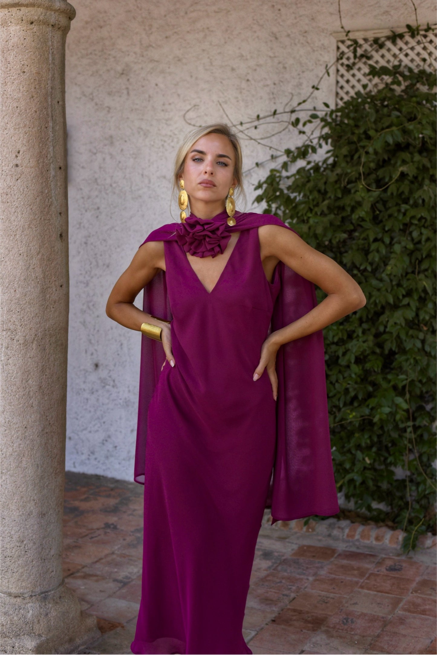 Vestido de Fiesta largo, lencero con escote de pico delante y en espalda. Con capita y broche de flor multiposición que da mucho juego y se puede colocar en el centro del escote o en un lateral.Confeccionado en gasa tono berenjena.